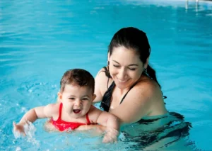Swim instructor giving early swim lessons for kids, by guiding an infant during a water safety progress milestone in a pool.